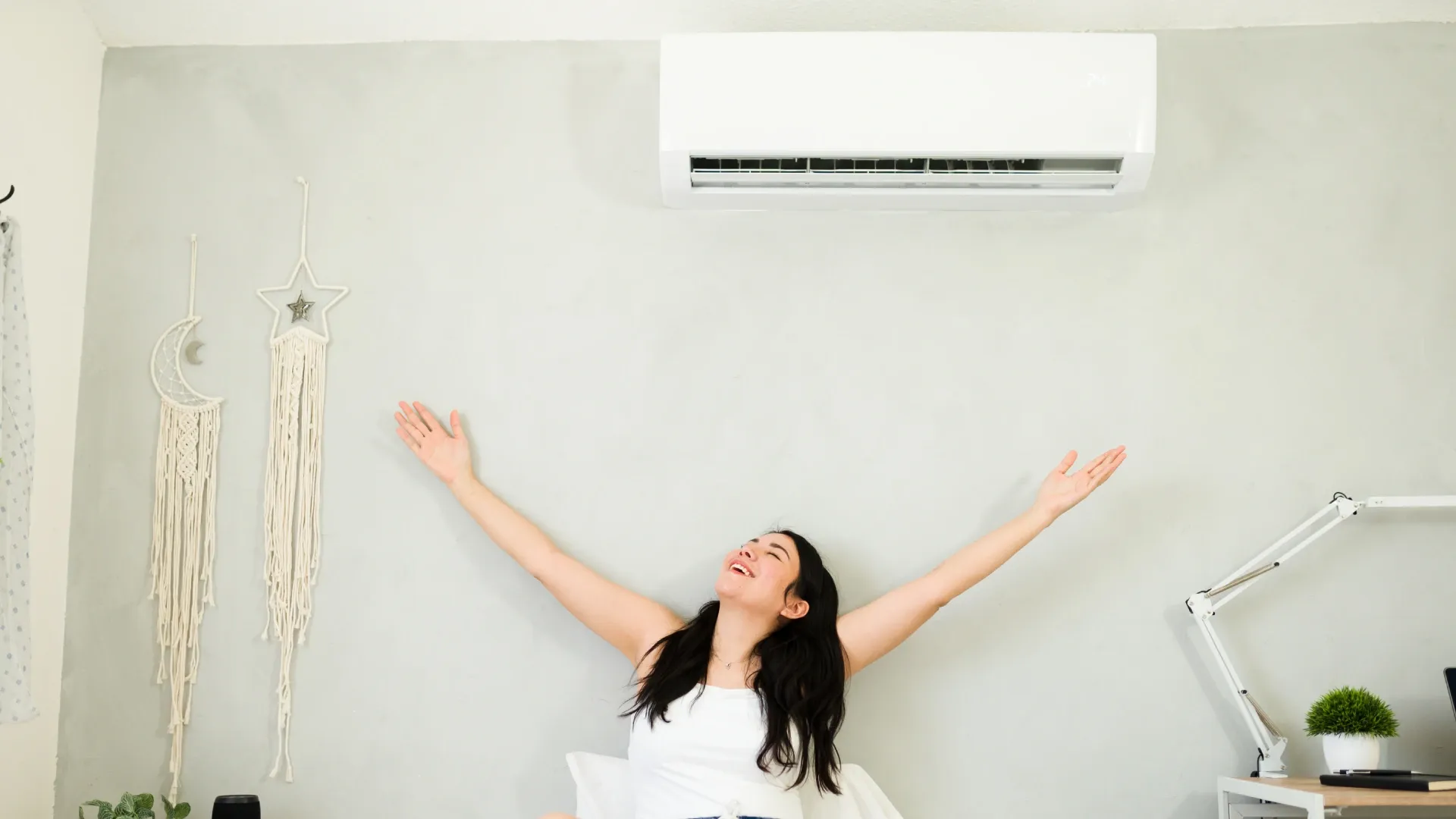 Jovem moça feliz sentada na cama com o ar-condicionado Split ligado 