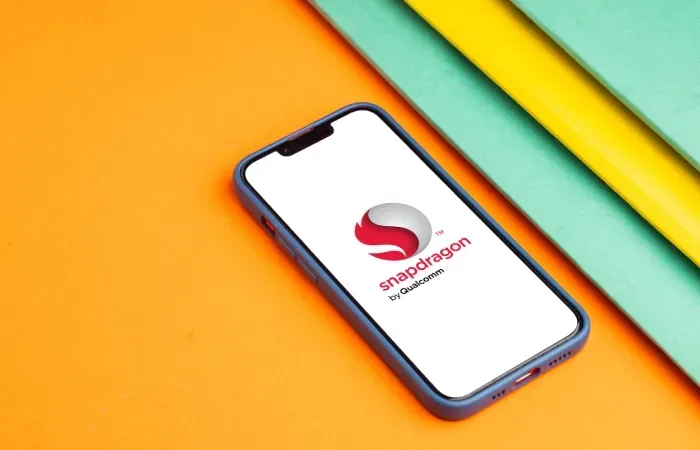 Smartphone com tela exibindo o logotipo do processador Snapdragon da Qualcomm, sobre superfície colorida.