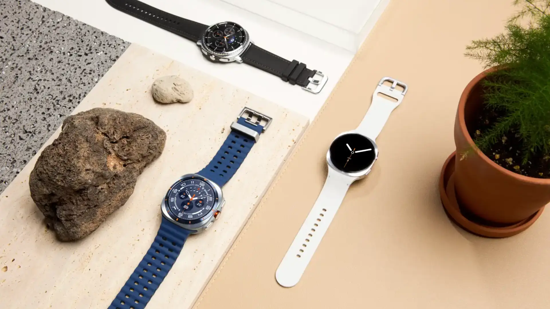 Três modelos de Galaxy Watch 8 Classic em cima de uma mesa ao lado de pedras e vaso de planta