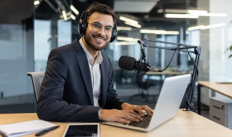 Homem sorridente de paletó trabalha em estúdio de locução com um Notebook 16Gb ao lado de microfone e tablet.
