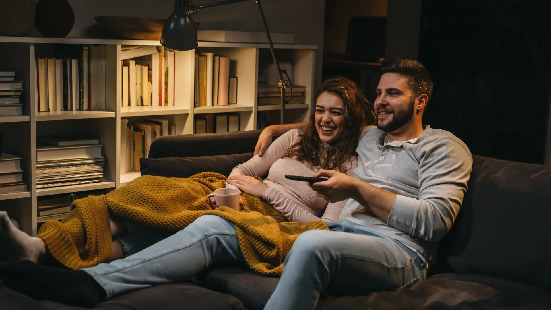 Casal sorridente assistindo TV juntos no sofá, cobertos com manta amarela, em ambiente aconchegante com estante de livros ao fundo