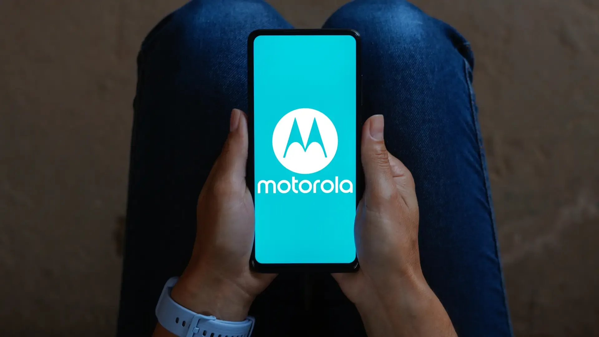 Uma pessoa está sentada com um celular. Na tela aparece a logo da Motorola