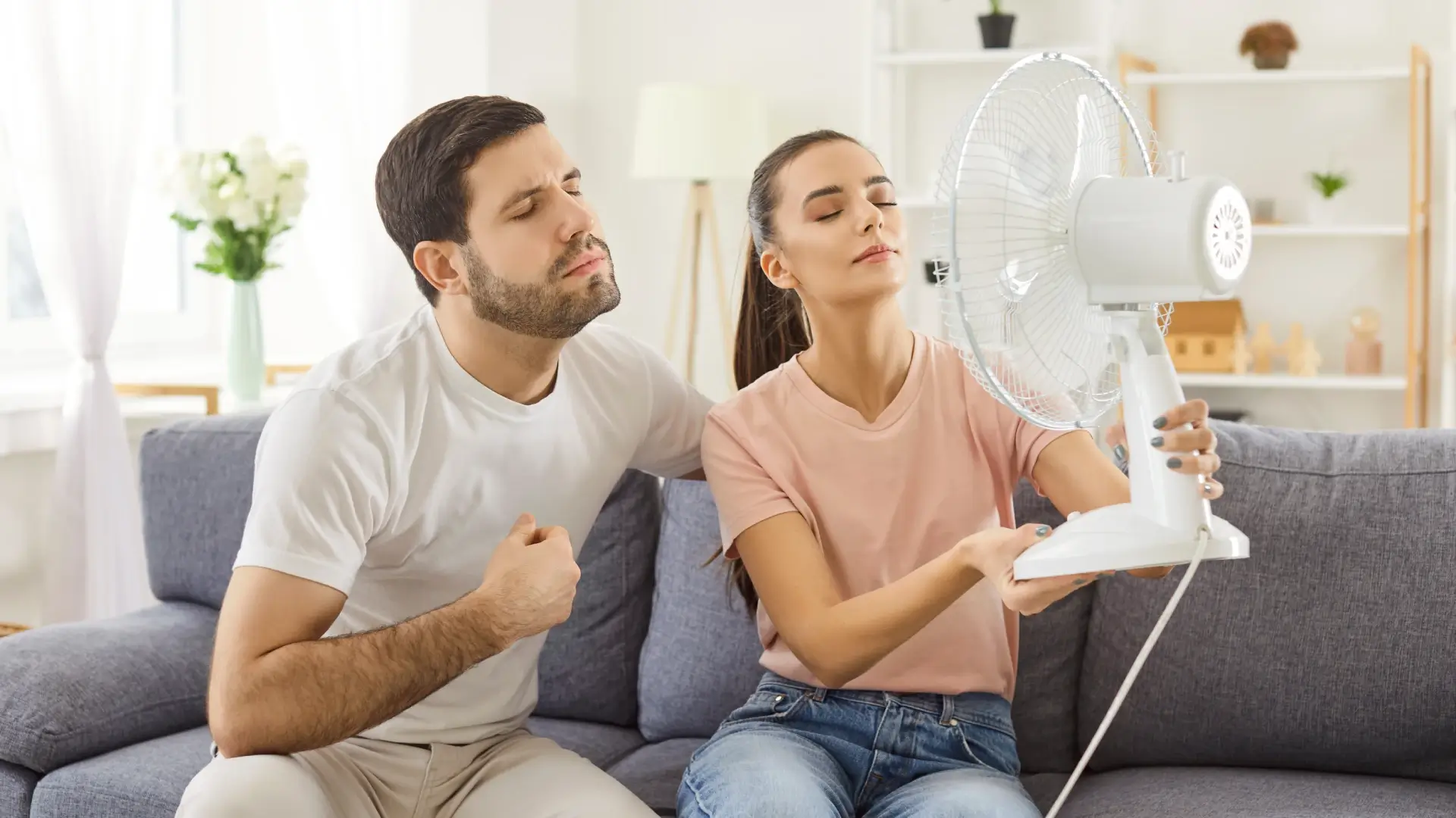 Casal sentado no sofá segurando o ventilador na direção deles 