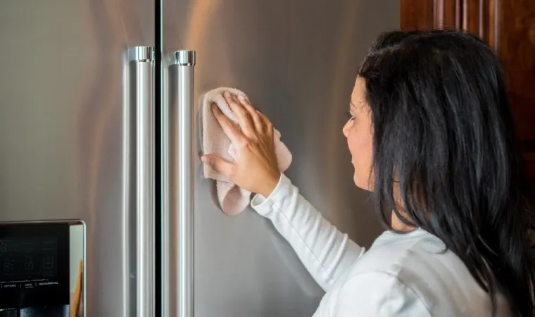 Mulher limpando uma geladeira de inox com pano microfibra.