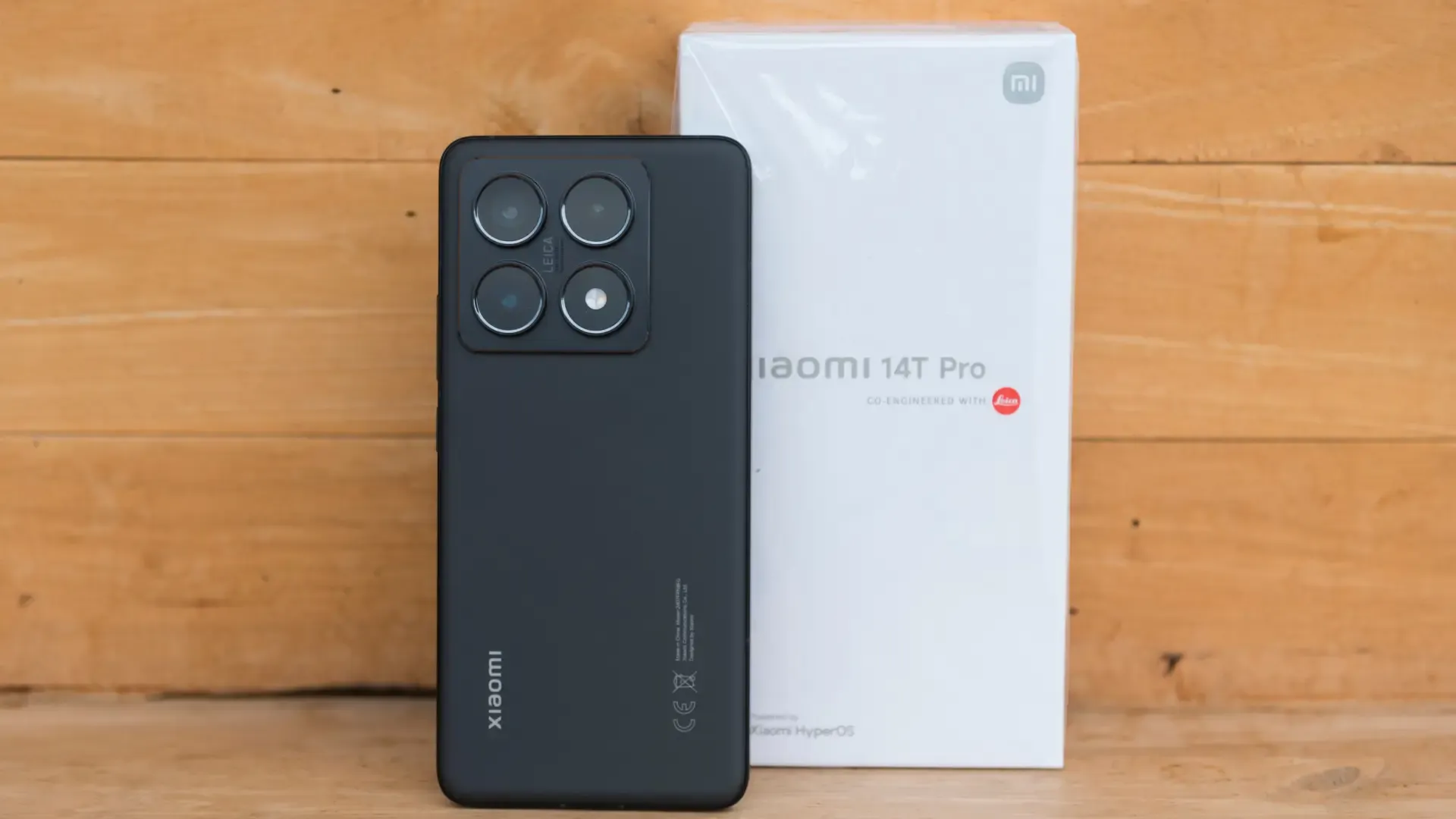 Um celular está em pé, ao lado da caixa, sobre uma estrutura de madeira