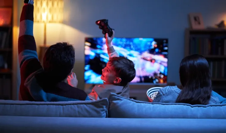 Imagem mostra três pessoas num sofá, sendo elas uma criança com um controle de videogame na mão comemorando