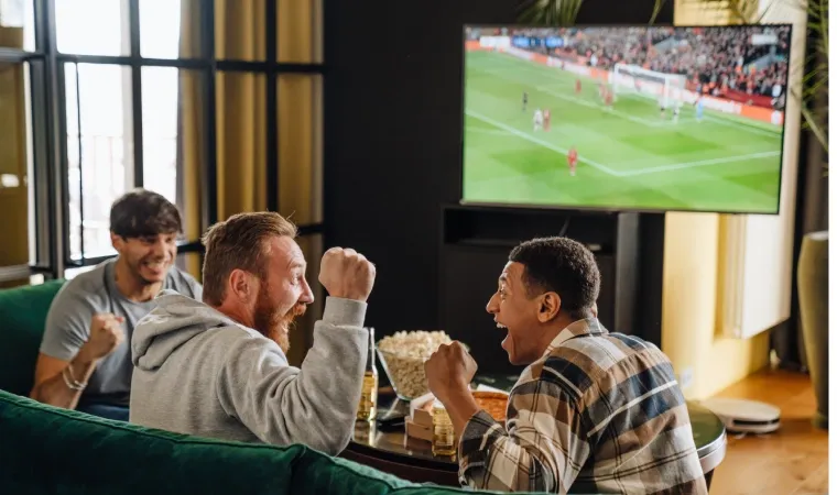 Imagem mostra três homens no sofá comendo pipoca e comemorando o resultado de um jogo de futebol.