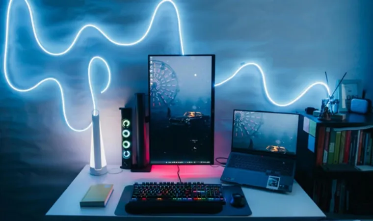 Foto de monitor vertical sobre uma mesa ao lado de notebook, junto de teclado e mouse e uma fita led emitindo luz azulada