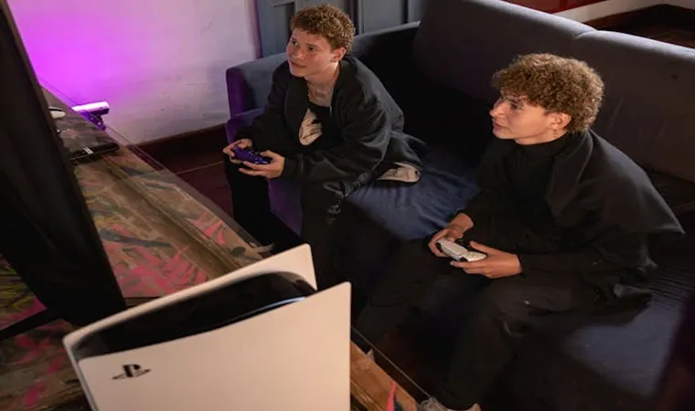 Foto com dois rapazes jogando um PlayStation 5 enquanto estão sentados no sofá