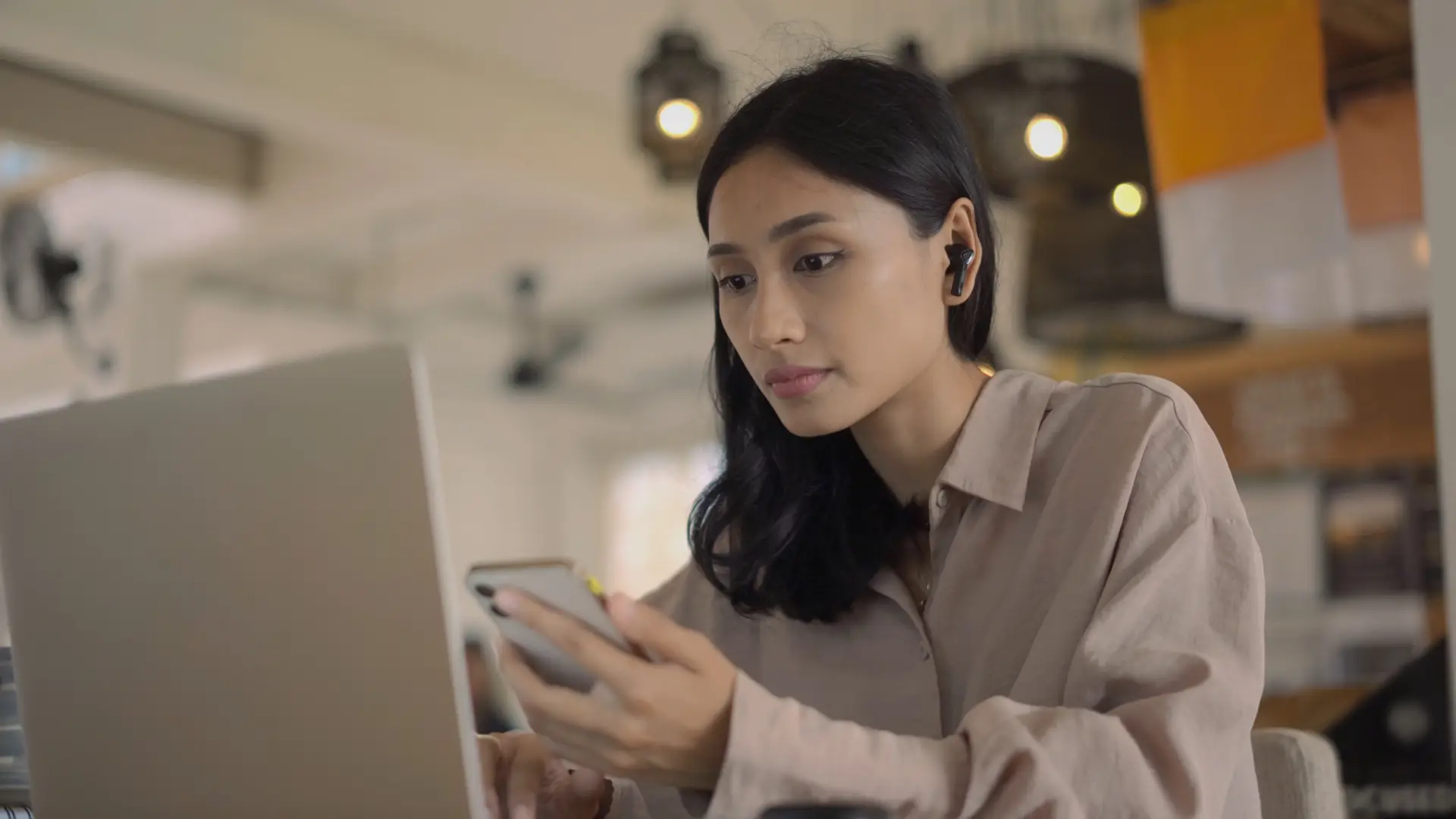 Uma mulher está sentada em frente a um notebook, usando fones de ouvindo e mexendo no celular