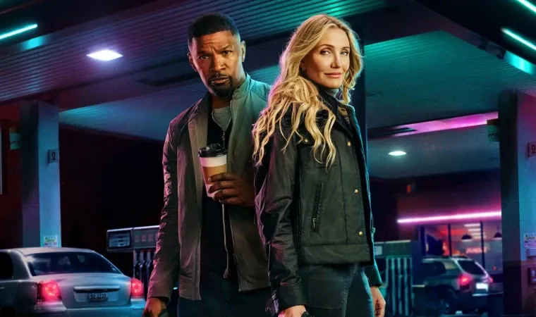 Cena do filme De Volta à Ação, mostrando um homem e uma mulher de jaquetas escuras em frente a um posto iluminado à noite, com expressão determinada.