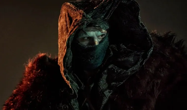 Cena do filme Frankenstein mostrando um homem encapuzado e coberto por um manto escuro, com expressão enigmática sob luz avermelhada. 