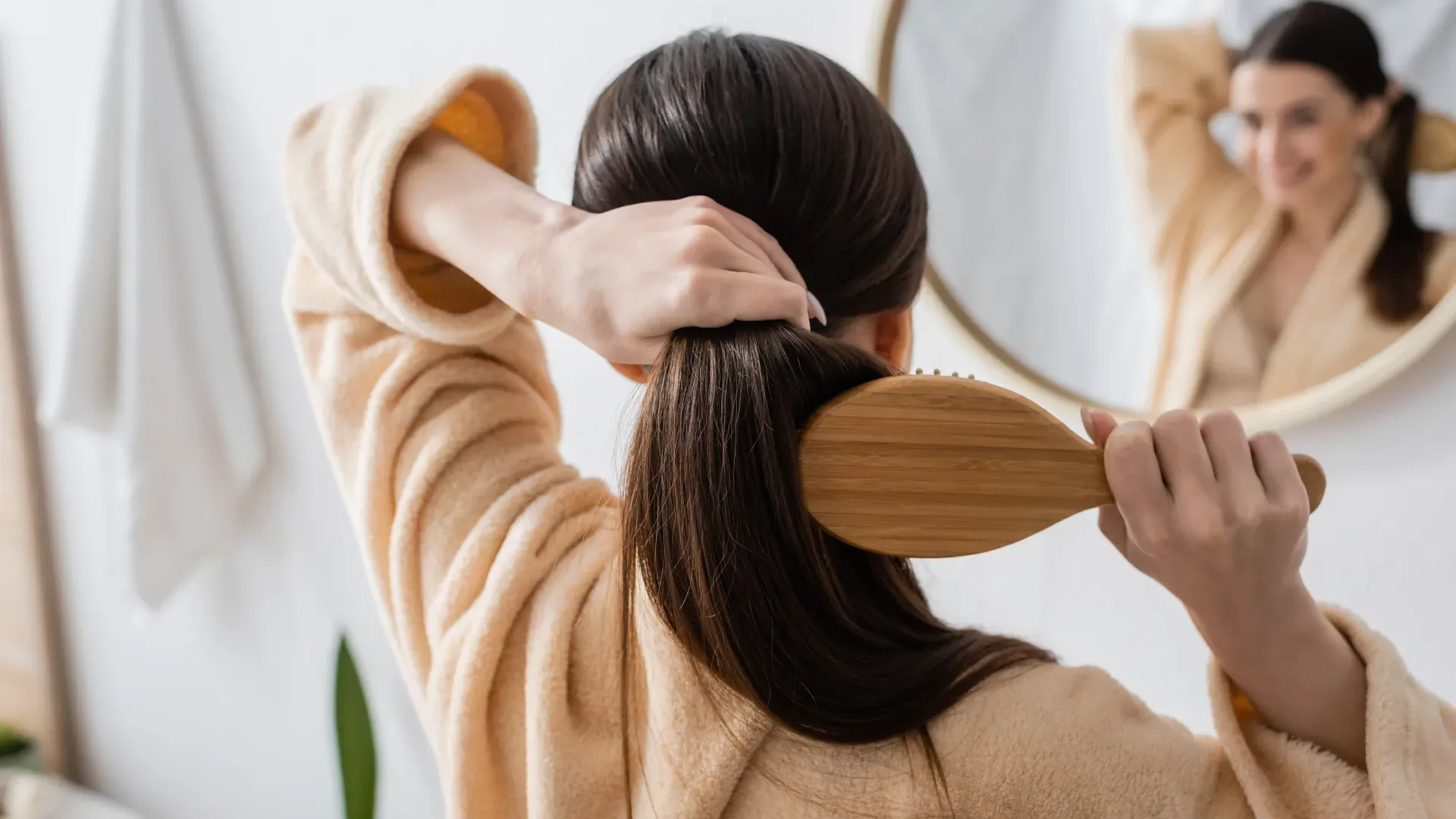 Mulher de cabelo escuro está de frente para um espelho penteando o cabelo