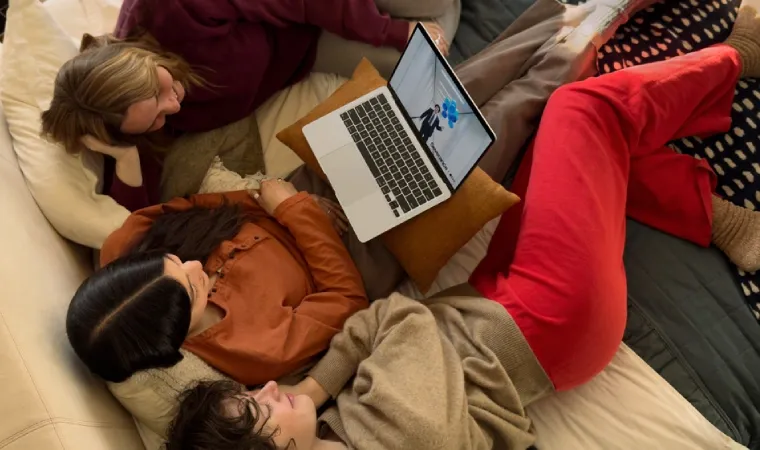 Imagem mostra três garotas deitadas em um sofá. A garota do meio tem um MacBook Air no colo.