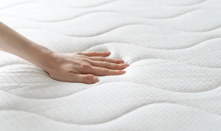 Mulher com cabelos cacheados, compridos e escuros, está vestindo um conjunto de calça e regata marrons. Ela está sentada em um colchão Ortobom Nanoelastic Pró-Saúde. No quarto vemos uma decoração minimalista, com mesinhas de cabeceira marrons, luminárias e livros