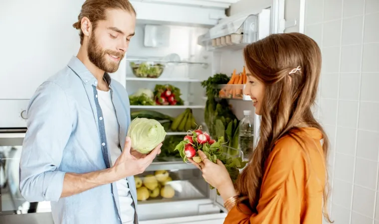 Casal olhando legumes com a porta da geladeira aberta