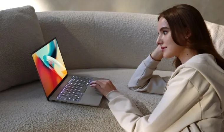 Imagem mostra uma moça utilizando um notebook com IA da ASUS. Ela está sentada no chão e o notebook está em cima de um sofá de cor clara.