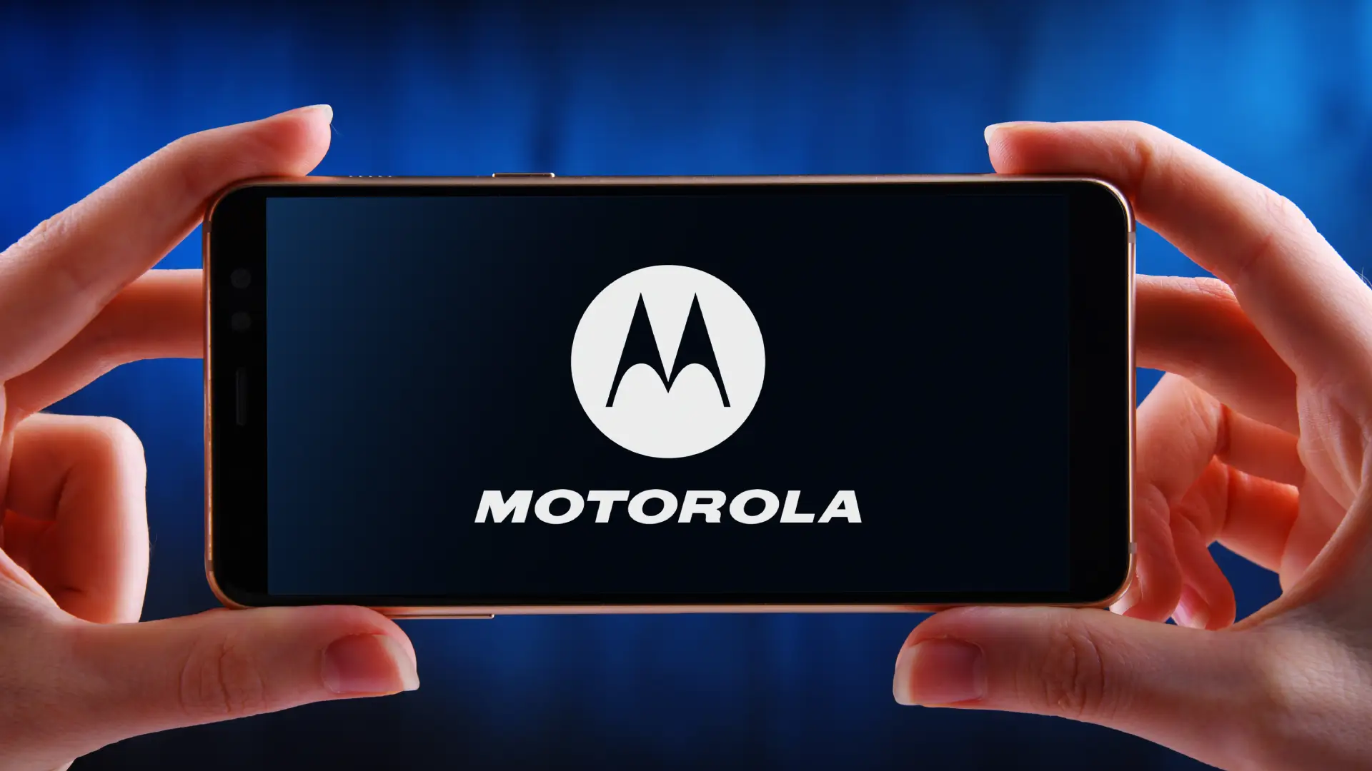 Um celular está na horizontal, sendo segurado por duas mãos. Na tela está escrito Motorola