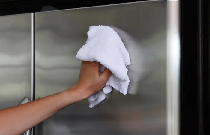 Mão limpando a porta de uma geladeira cinza com pano branco.