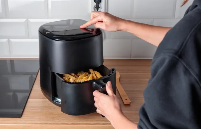 Pessoa abrindo air fryer preta com cesto de batatas fritas na cozinha.