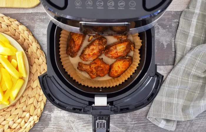 Asas de frango assando em airfryer aberta sobre bancada com batatas ao lado.