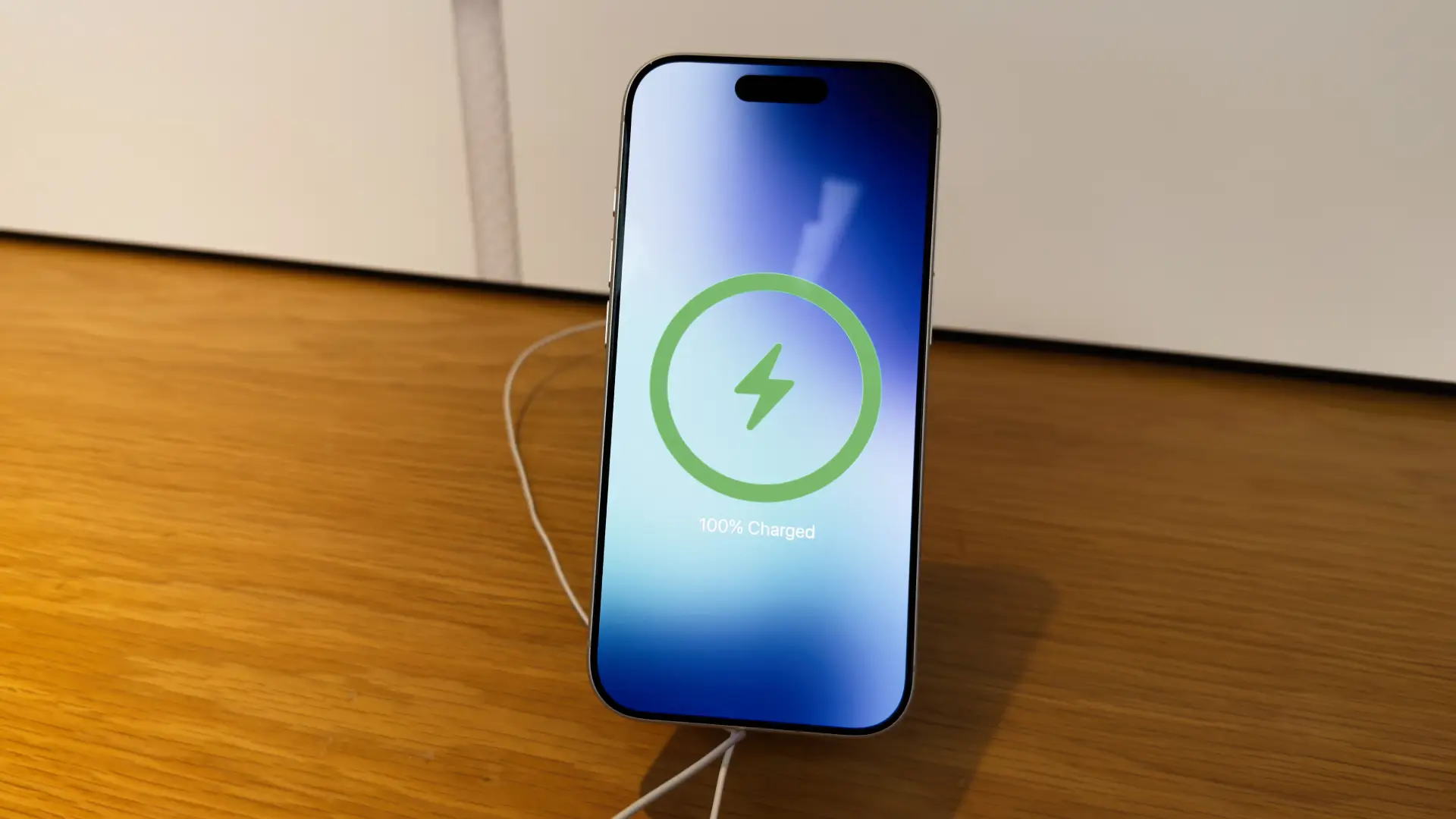 Imagem de um iPhone 17 Pro apoiado sobre uma mesa de madeira enquanto carrega sem fio, com indicador de bateria cheia exibido na tela.