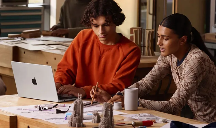 Imagem de dois estudantes de arquitetura usando um Macbook Pro com processador M5 sobre uma mesa com desenhos e pequenas maquetes de prédios
