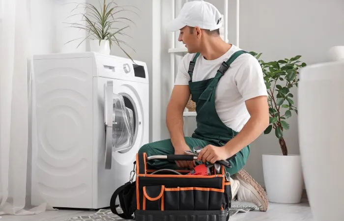 Técnico ajoelhado ao lado de uma máquina de lavar, com bolsa de ferramentas aberta, realizando manutenção preventiva.