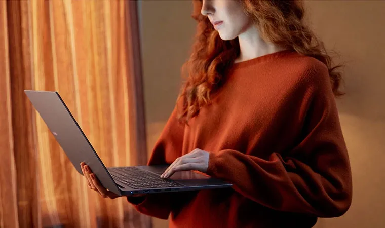 Foto de uma mulher ruiva em pé segurando com uma das mãos um notebook Samsung Galaxy Book4 aberto enquanto navega com a outra mão.