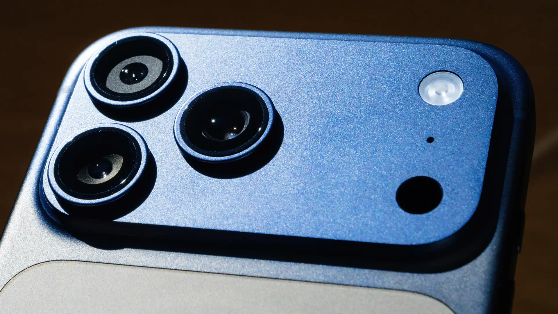 Imagem em close do módulo traseiro de câmeras de um iPhone 17, destacando três lentes e o acabamento metálico ao redor do conjunto.