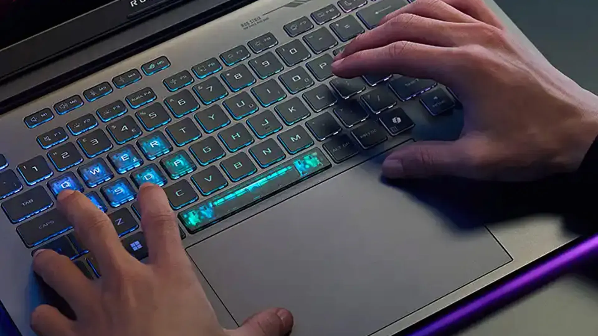 Teclado iluminado do ASUS ROG Strix G18 (2026) com RGB azul ativo e barra de luz roxa visível na lateral do chassi
