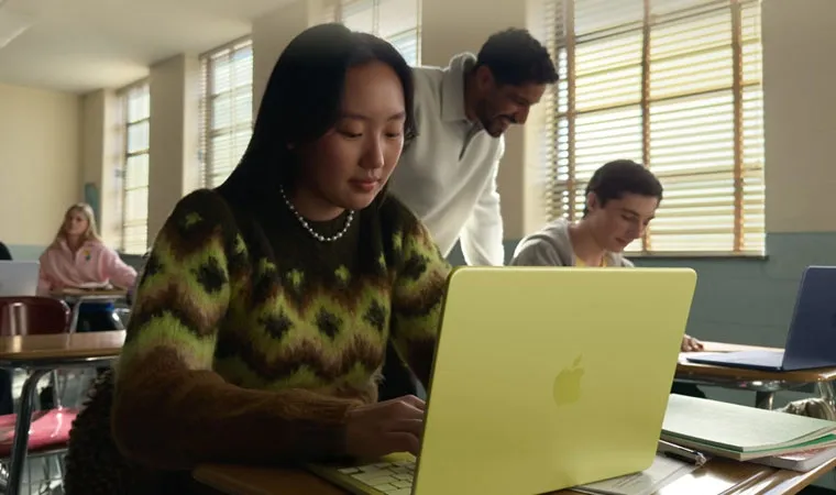 Foto de uma mulher jovem usando o MacBook Neo para estudar em uma sala de aula enquanto um professor ajuda outro aluno ao fundo