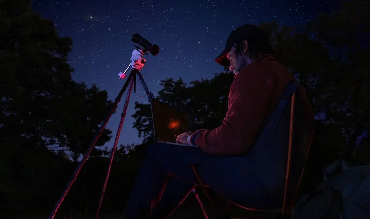 Imagem de um astrônomo usando um MacBook Pro no colo em meio à natureza enquanto aponta um telescópio para o céu