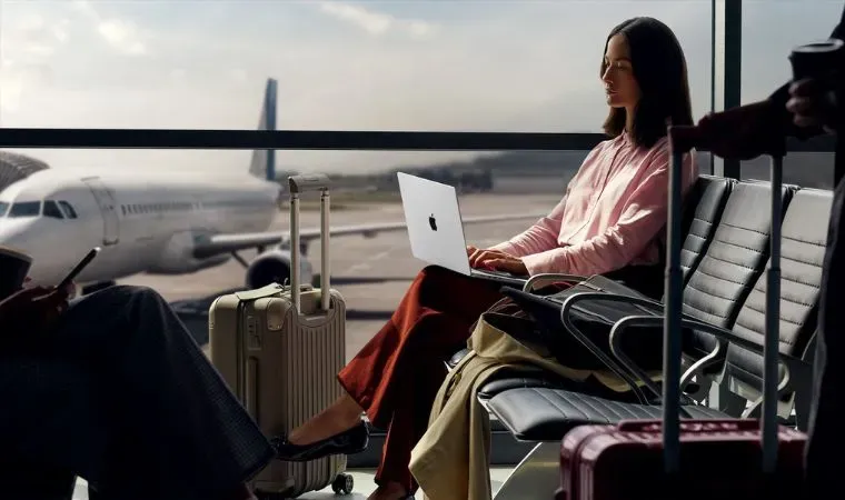 Mulher sentada em um aeroporto com um avião ao fundo trabalha usando um Apple Macbook Pro M5