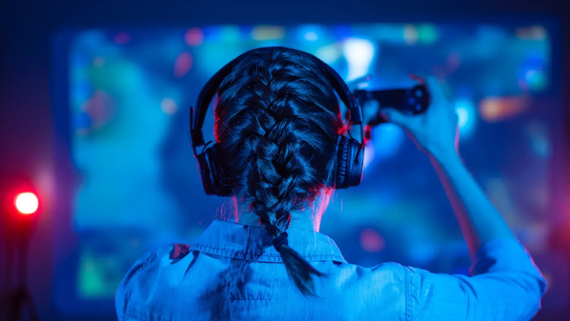 Mulher de costas em frente a uma TV de tela grande, usando headphone e com controle de videogame nas mãos