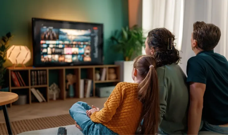 Família composta por homem, mulher e menina assiste TV no ambiente confortável da sala de casa.