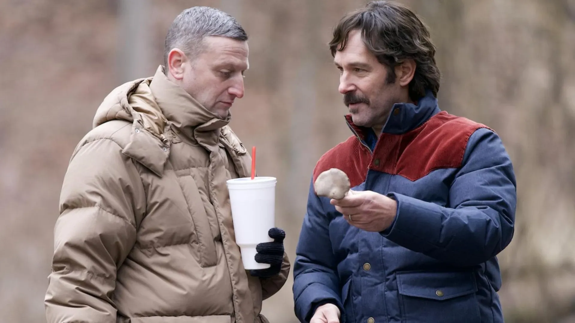 Dois homens de casaco de inverno em um parque, um segurando um copo e o outro um cogumelo, em cena do filme Friendship
