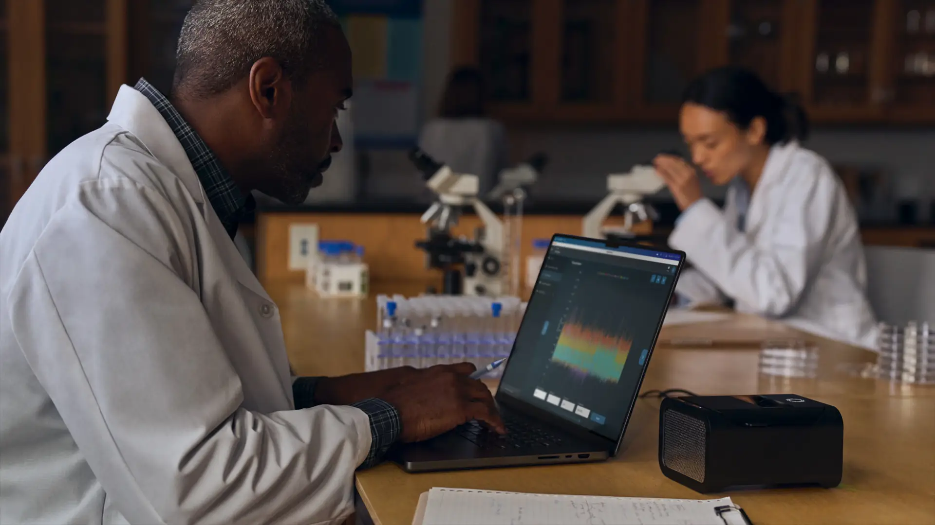 Cientistas em um laboratório com microscópio e ampolas trabalham com gráficos em um notebook Apple Macbook Pro M4