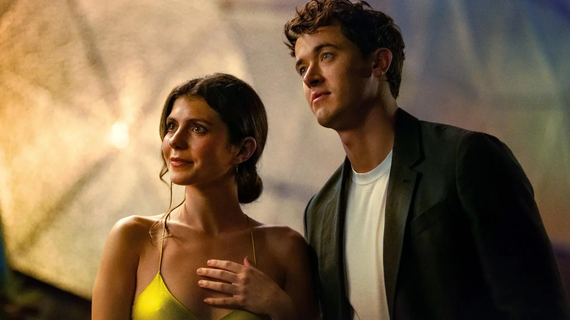 Homem e mulher jovens lado a lado em ambiente iluminado, ela com vestido amarelo e ele de blazer escuro, em cena do filme De Férias com Você