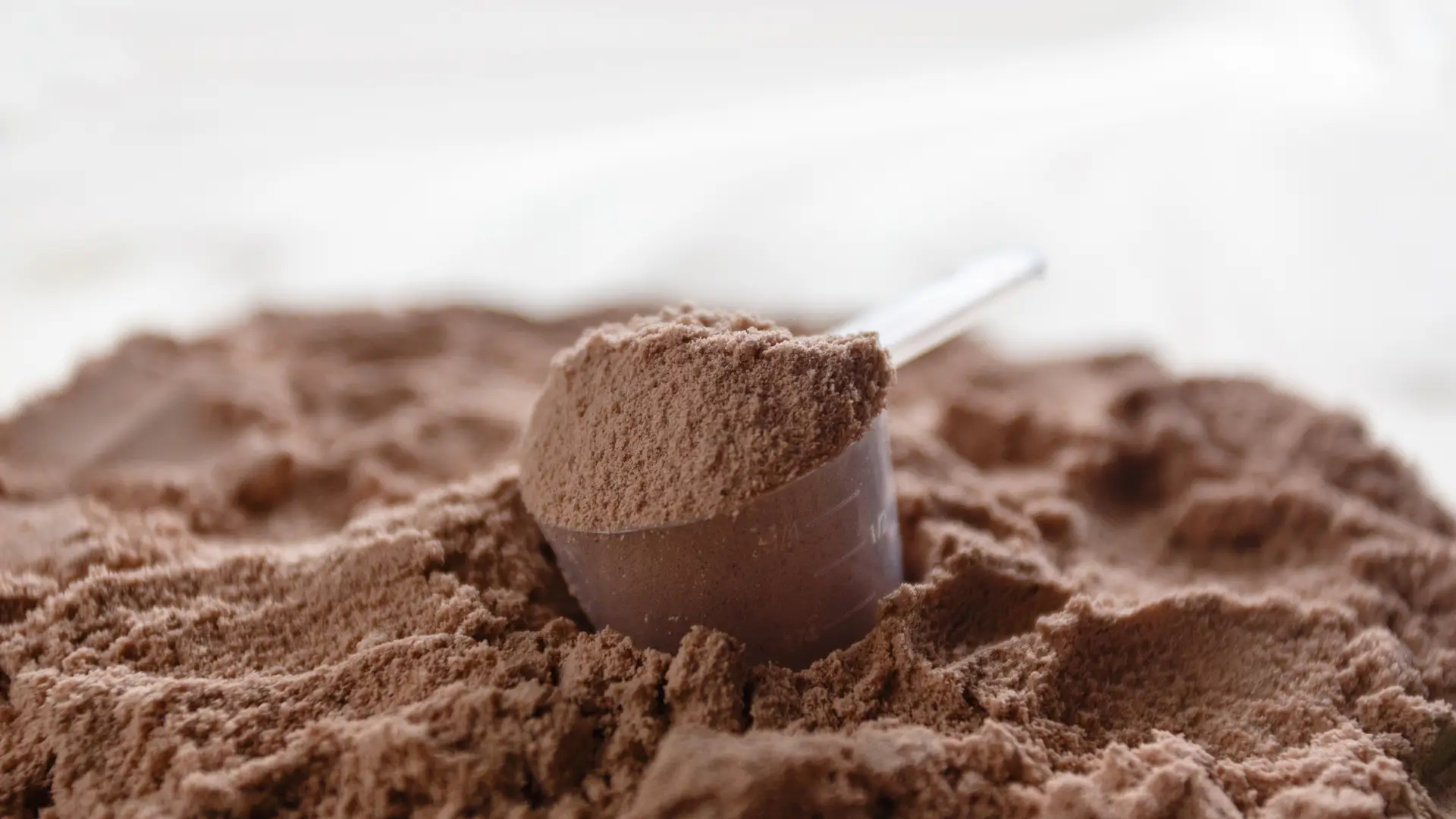 Colher medidora apoiada sobre whey protein em pó sabor chocolate em fundo de madeira