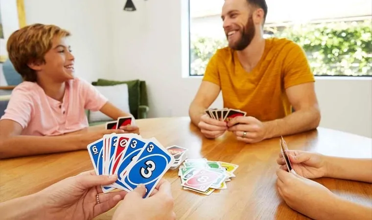 Família jogando UNO vista em primeira pessoa com uma mãe olhando para suas cartas coloridas em mãos enquanto o pai e filho riem à sua frente 