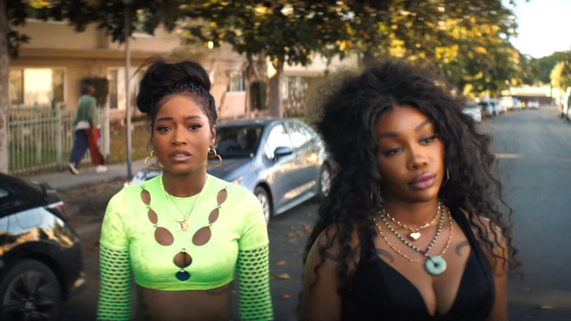 Duas mulheres jovens em expressão séria numa rua de bairro residencial, uma com blusa verde neon e outra de top preto, em cena do filme One of Them Days