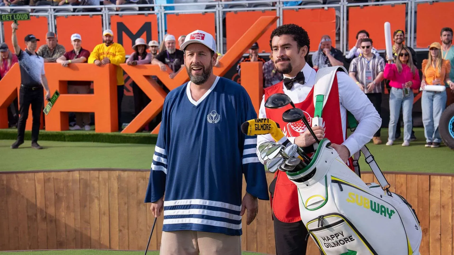 Adam Sandler com tacos de golfe e camiseta de hóquei ao lado de um caddie num campo de golfe lotado de torcida, em cena de Um Maluco no Golfe 2