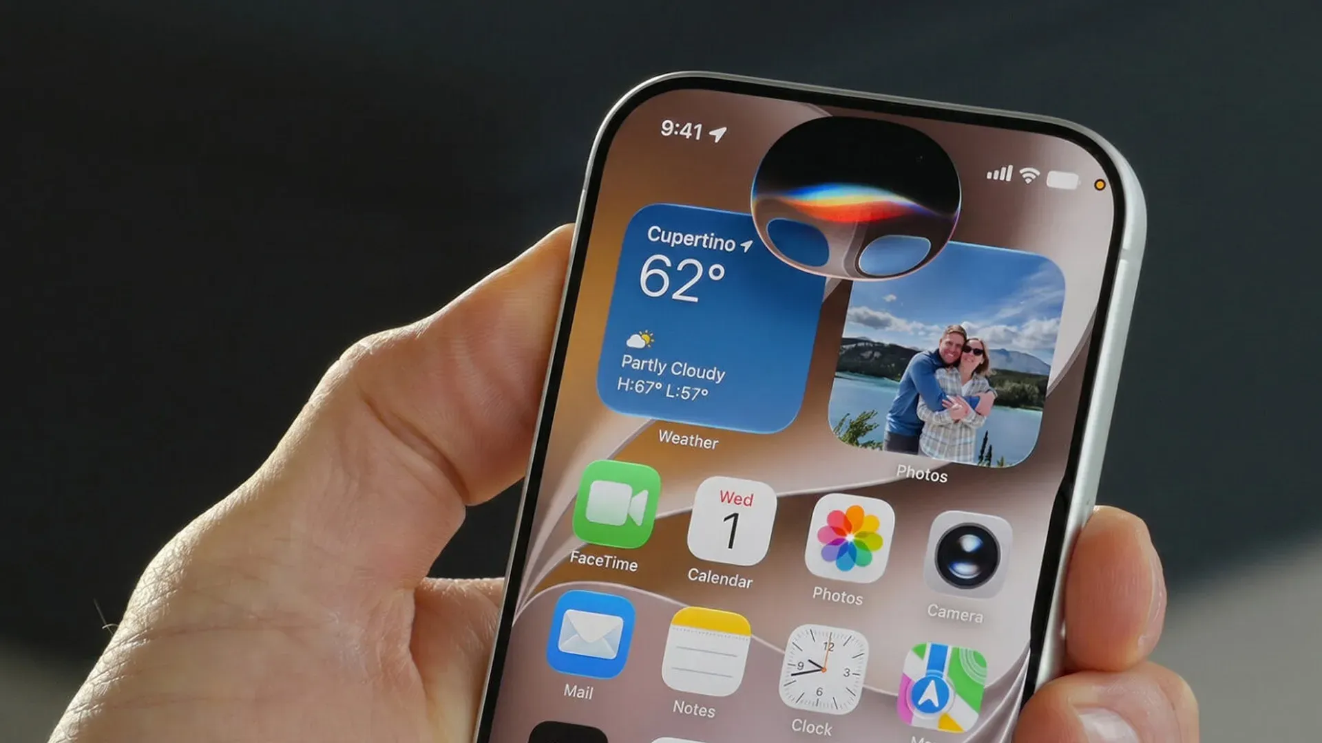 Mão segurando iPhone com a Siri AI exibida no Dynamic Island, sobre a tela inicial com widgets de clima e fotos 