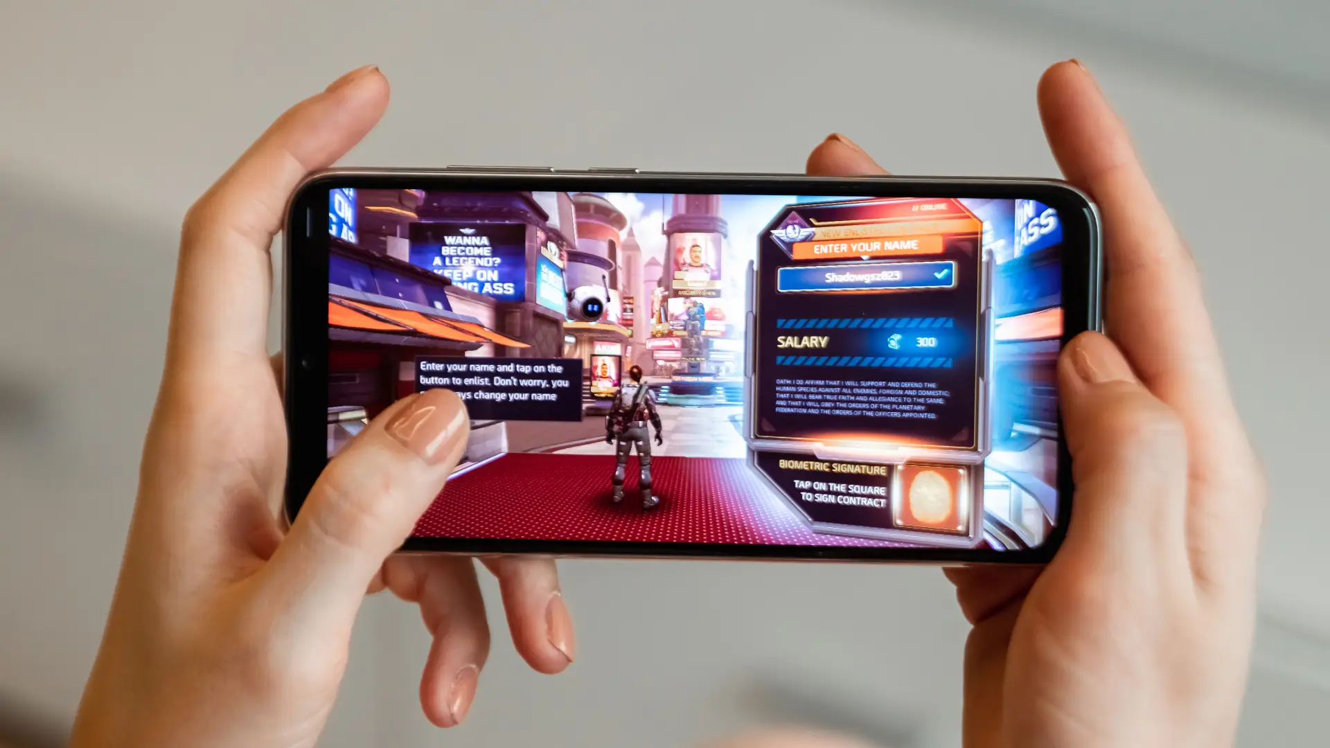 Mãos seguram smartphone na horizontal exibindo jogo futurista com personagem e painel de cadastro.