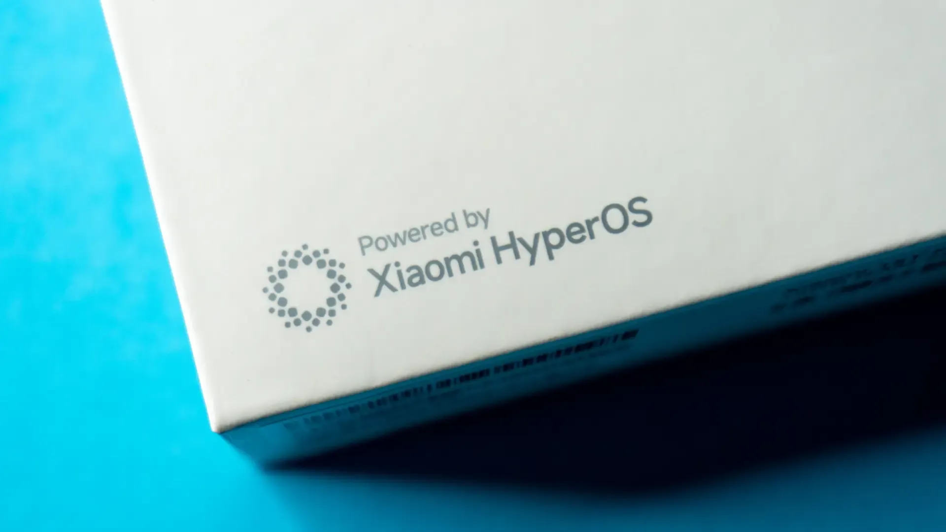 Close-up de caixa branca com inscrição “Powered by Xiaomi HyperOS” sobre fundo azul.