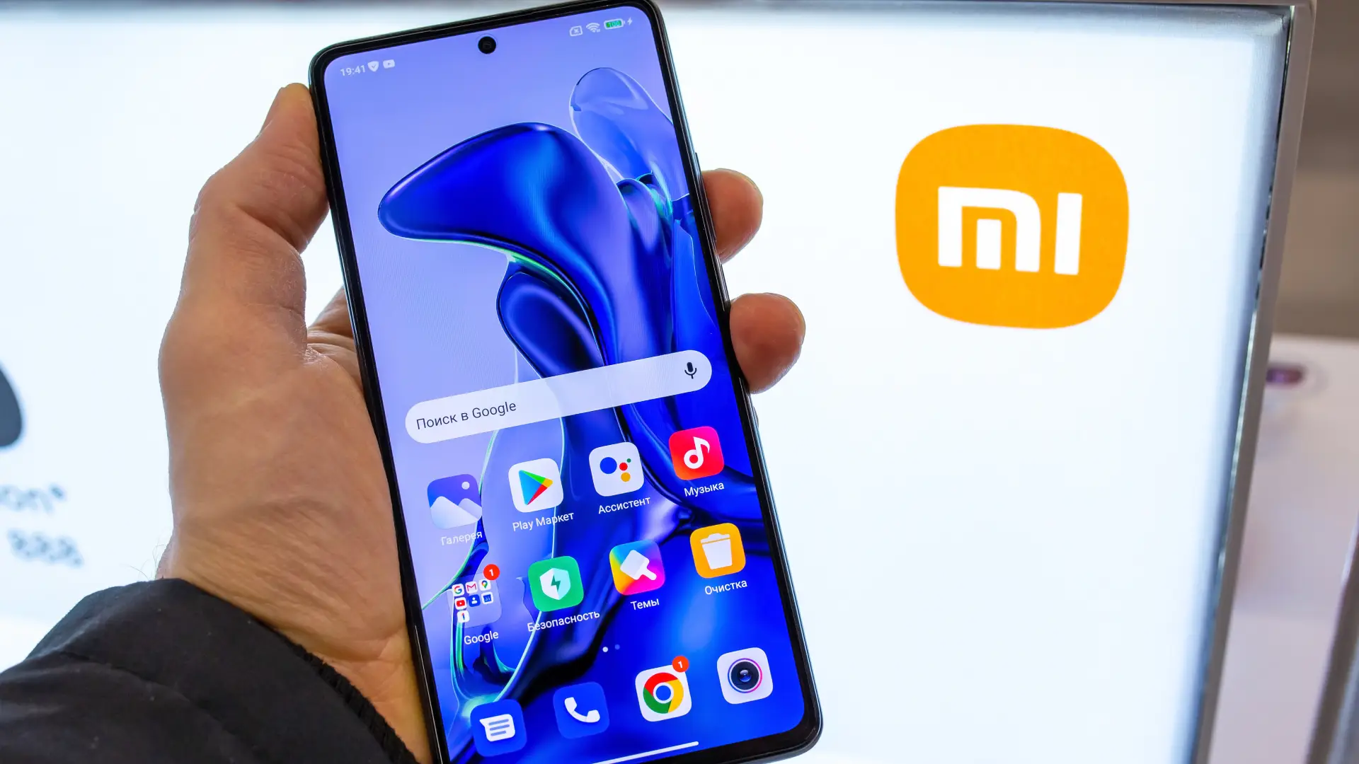 Pessoa segura smartphone Xiaomi com tela inicial azul diante de painel com logo mi.