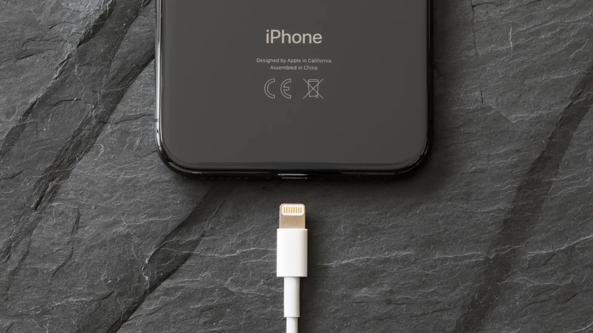 Parte traseira de iPhone preto próxima a um cabo Lightning sobre superfície de pedra.